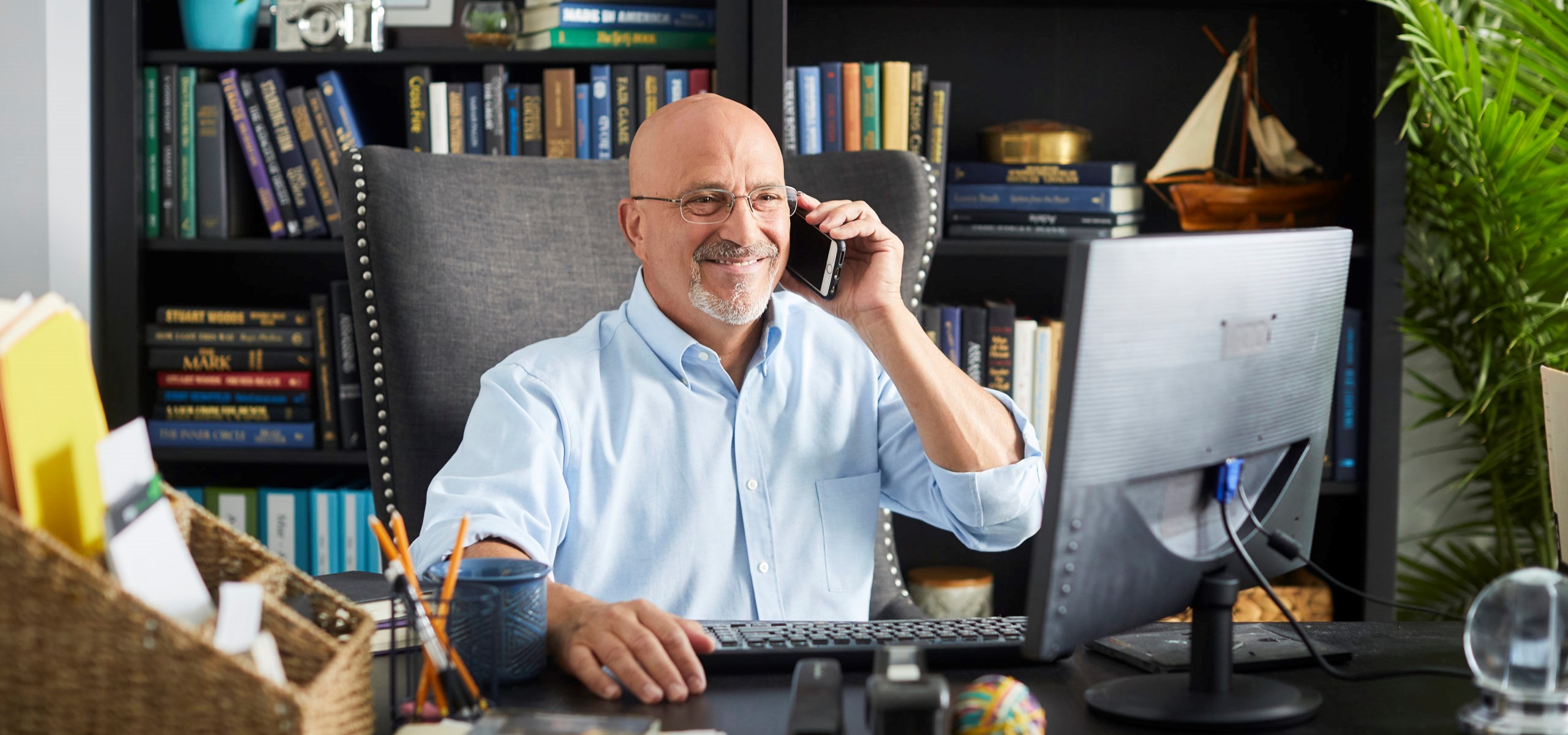 Man in his 60s smiling takes a phone call in front of his computer monitor
