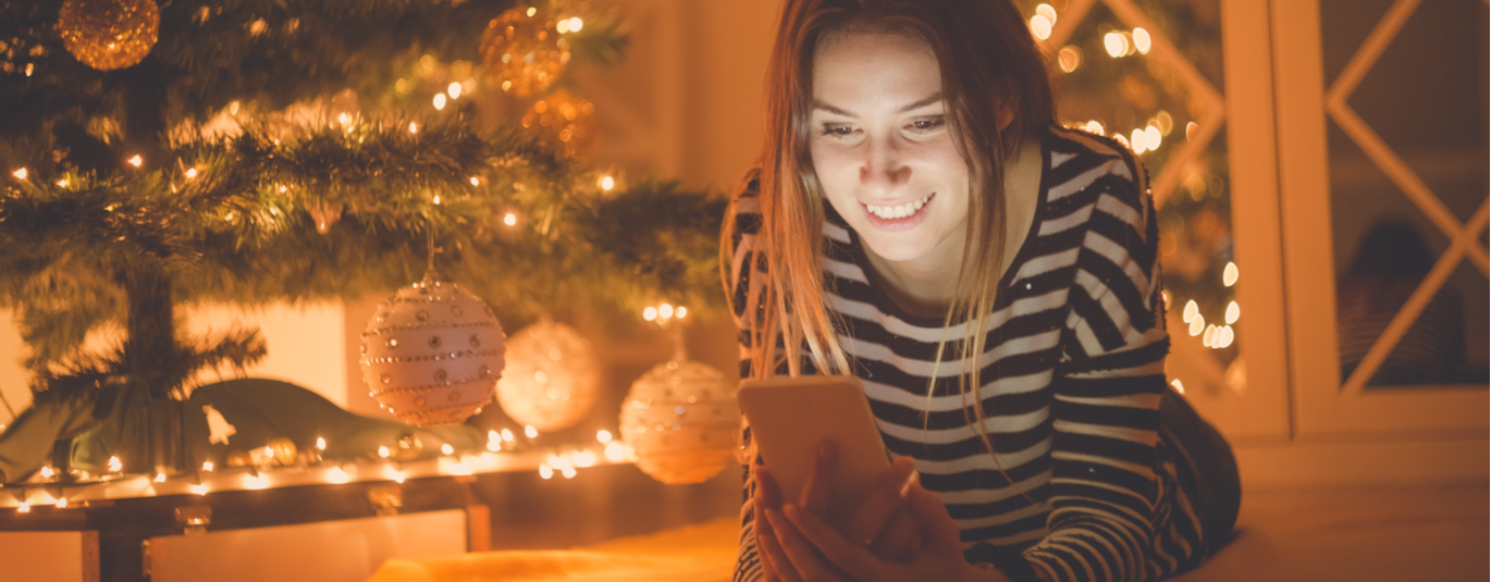 Happy woman lying on the floor and looking at mobile phone screen. Christmas tree with decoration is next to her.