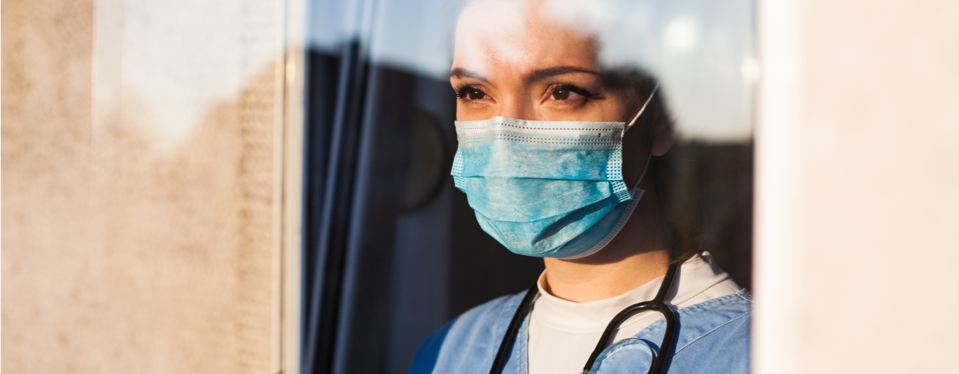 Female healthcare worker in scrubs a nd masks looks out the window with a determined expression