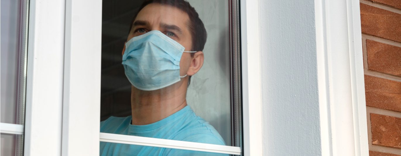 Man in a medical mask looks out the window of his home, where he is quarantined.