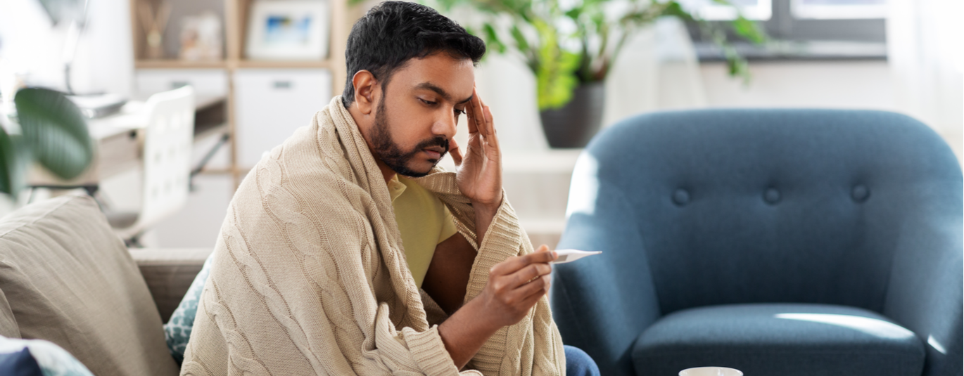 Man sick at home under a blanket, looking at a thermometer